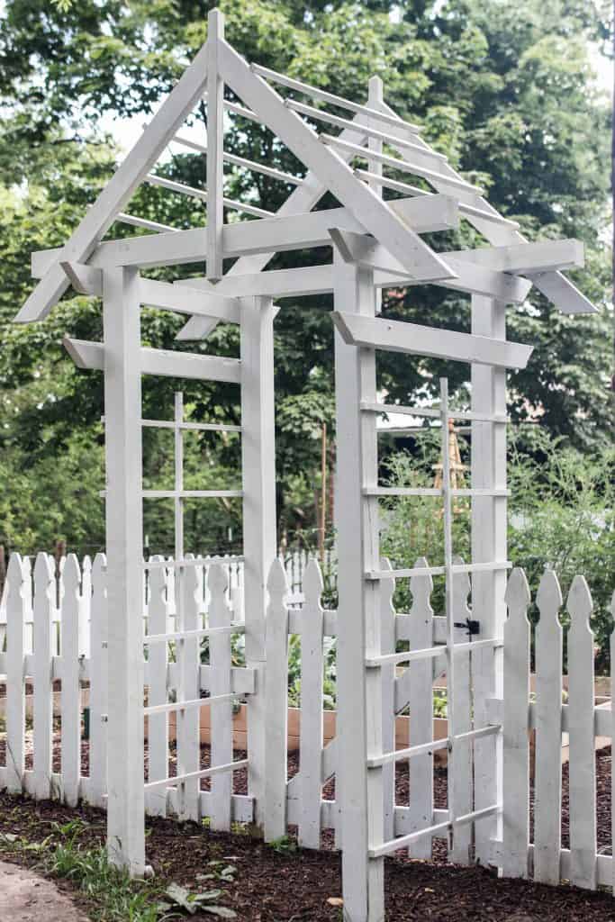 side view of a white arbor against a white picket fence with trees in the background