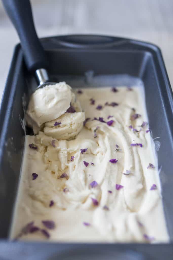ice cream scoop in a bread loaf pan full of lavender ice cream with fresh lavender flowers sprinkled on top.