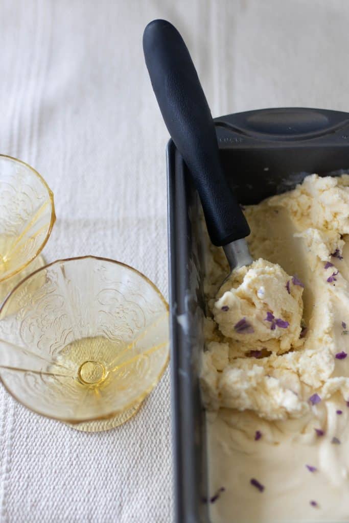 lavender ice cream in a loaf pan with an ice cream scoop. Two glass Sunday glasses to the left