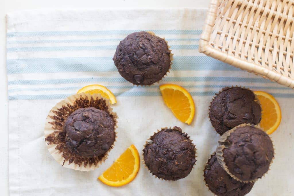 overhead photo of zucchini chocolate muffins on a blue and white stripped table cloth with orange slices spread about the muffins