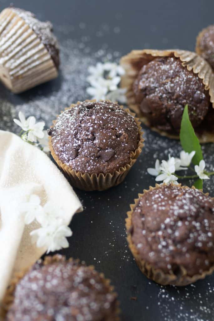 5 sourdough chocolate zucchini muffins on a black countertop with white flowers spread throughout
