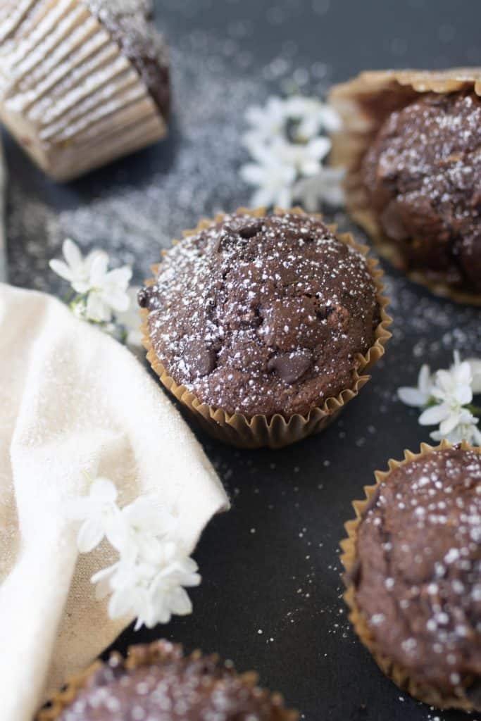 five sourdough chocolate zucchini muffins on a black countertop with flours