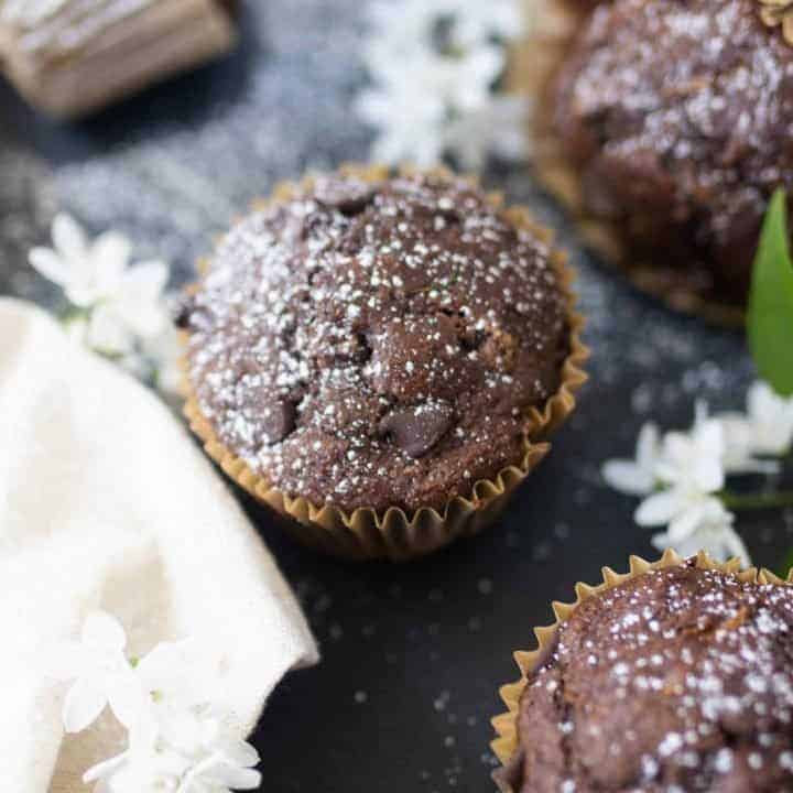 sourdough chocolate zucchini muffins spread out on a black countertop with white flowers and a white napkin on the countertop