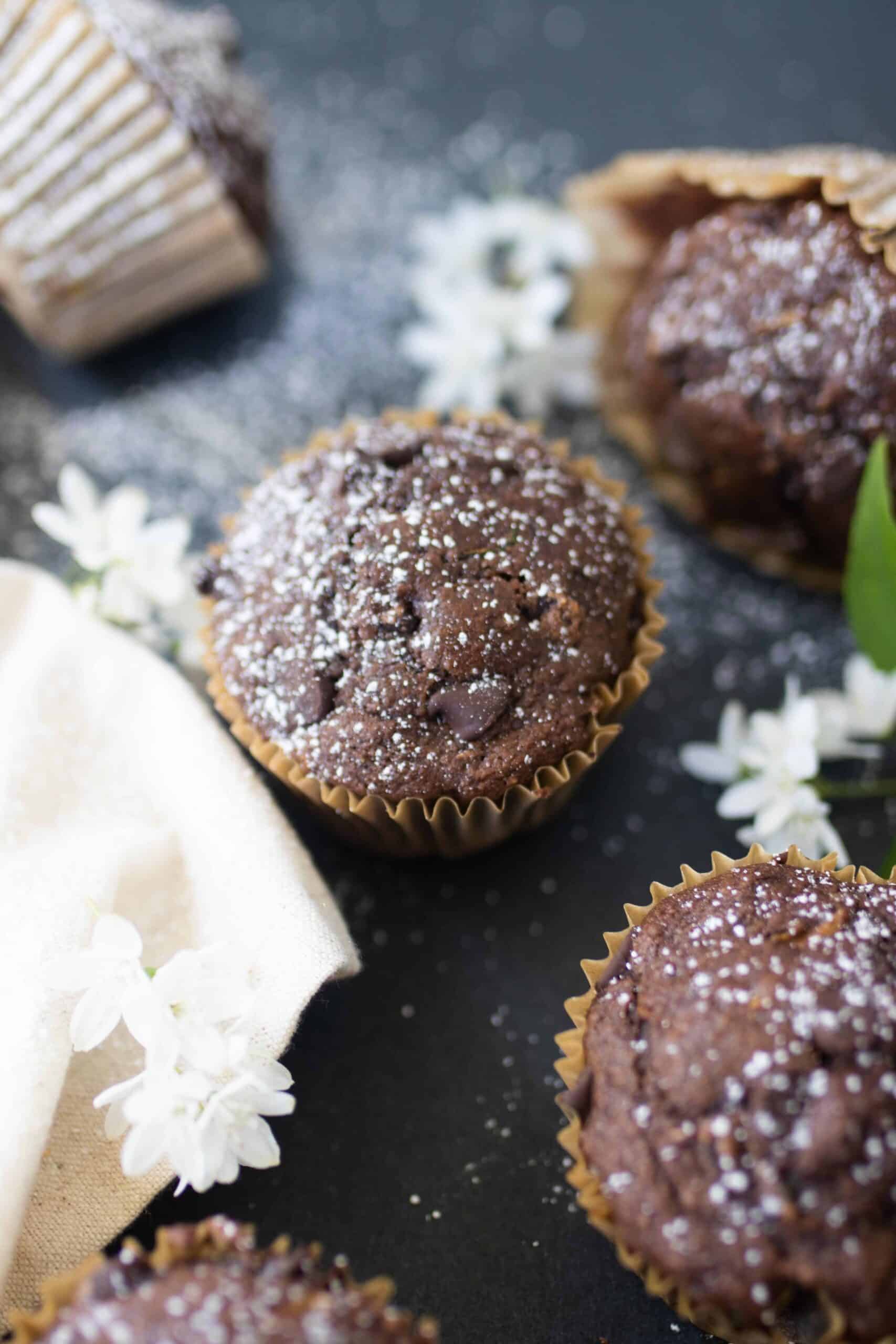 sourdough chocolate zucchini muffins spread out on a black countertop with white flowers and a white napkin on the countertop