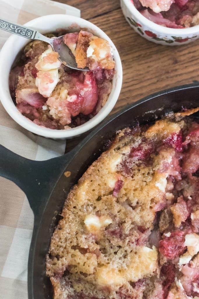 sourdough strawberry cream cheese cobbler in a cast iron skillet with a portion in an antique cup with a spoon in the back