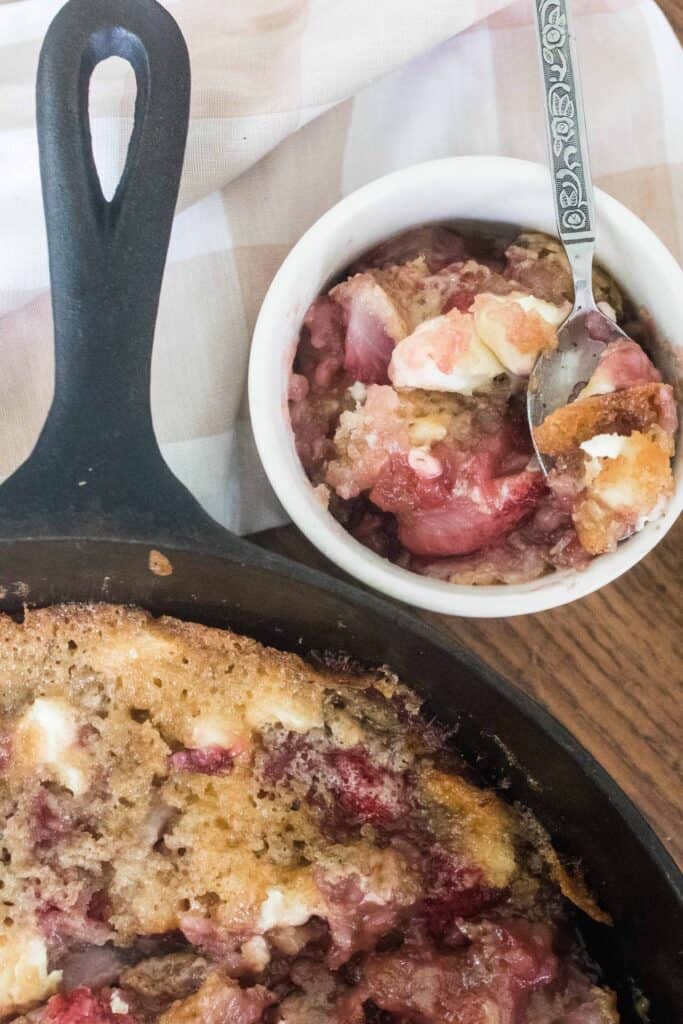 sourdough strawberries and cream in a cast iron skillet with a white ramekin of cobbler with a fork behind it