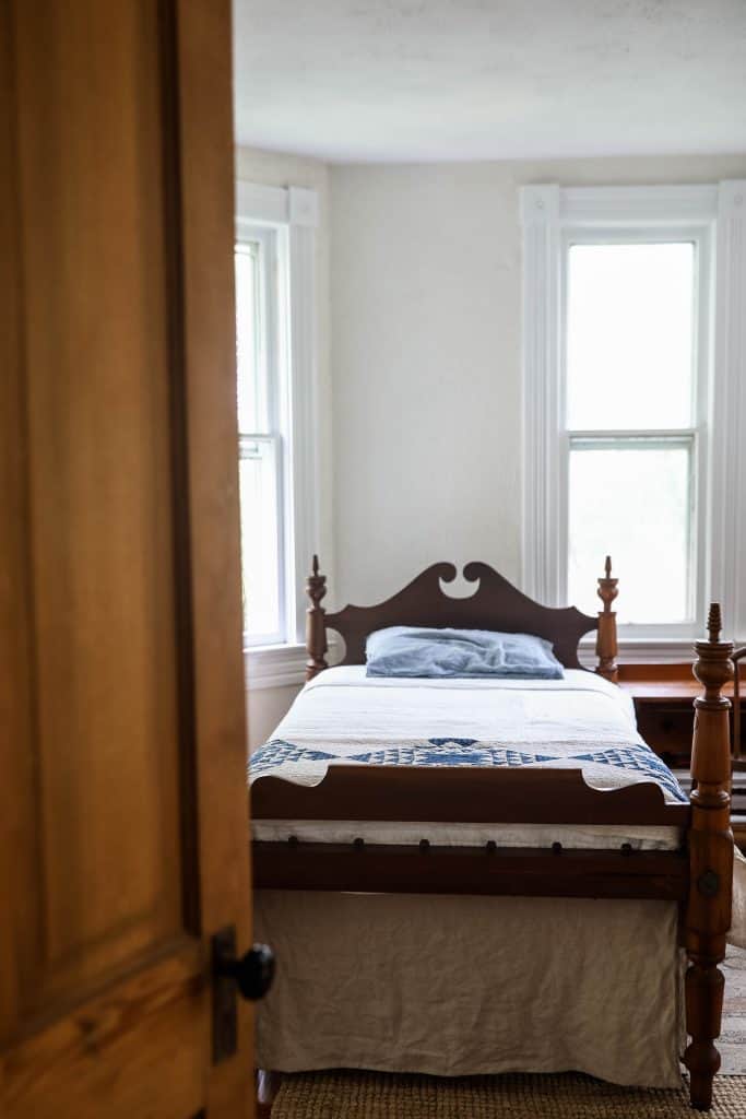 front view of an antique bed with a white bedspread, blue and white quilt at the foot, and a blue pillow near the headboard