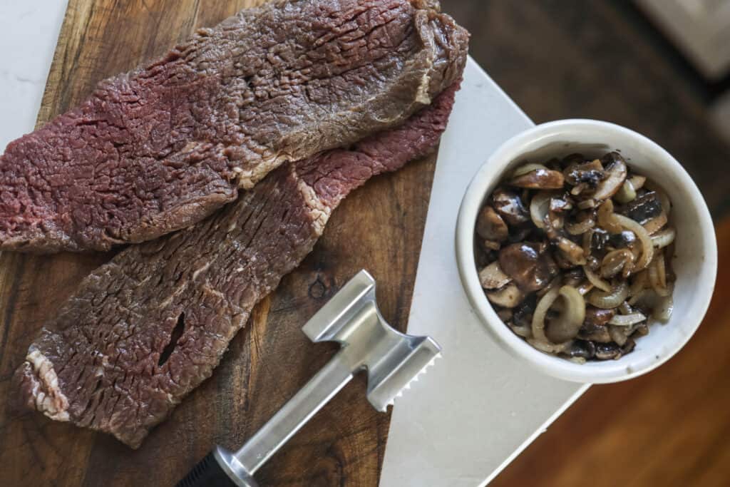 round steak pounded with meat mallet for tenderization, a bowl of caramelized onions sit to the right