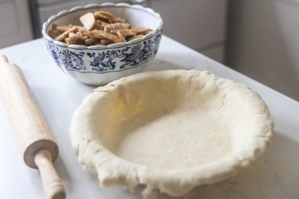 pie crust in a pie dish with sliced spiced apples in a bowl in the background