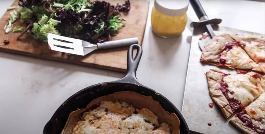 sourdough pizza in a cast iron skillet and another to the right on a pizza stone with salad greens on a cutting board to the left