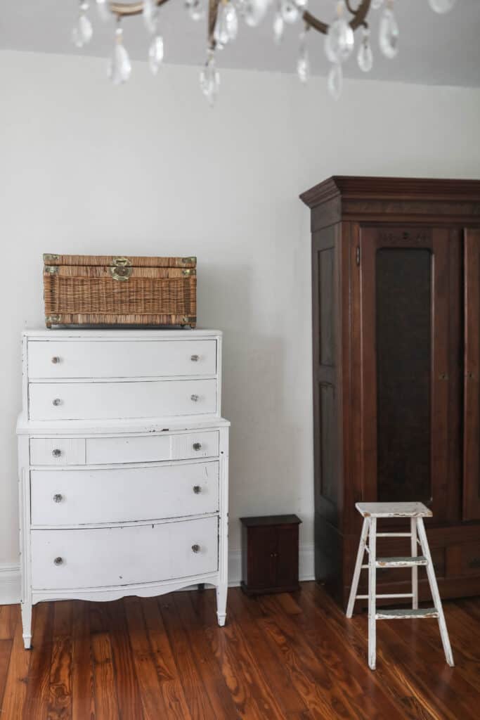 white antique dresser with a wicker chest on top. To the right in a wood armoire