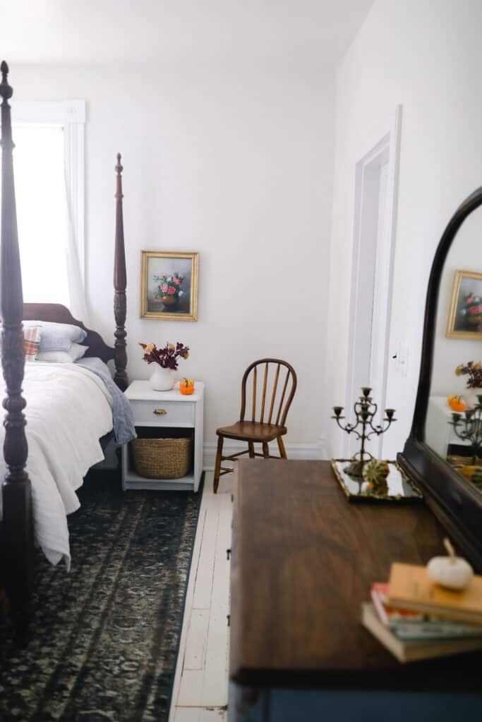 orange and brown books stacked on a dresser with a small white pumpkin on top. Fall florals in a white vase and a pumpkin on a night stand and fall linens on a antique bed.