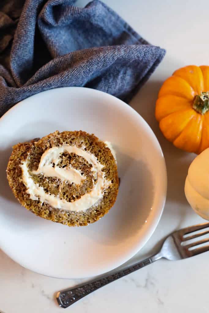 one slice of a pumpkin roll with cream cheese filling on a white plate with two small pumpkins to the right and a blue napkin in the background