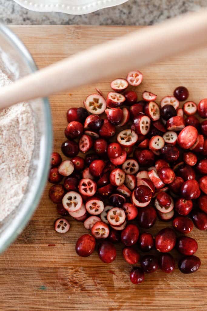 sliced cranberries on a cutting board with a bowl of dry ingredients to the left