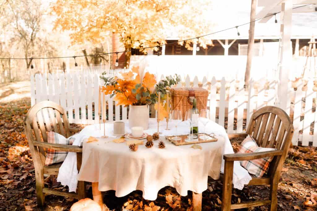 wooden table with tan linen table cloth with a crock of dried leaves and eucalyptus with a picnic basket with a barn with a picket fence in the background