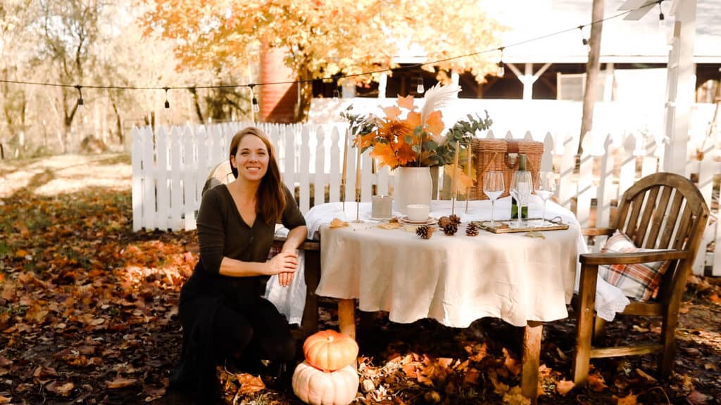 women sitting at a table set for a fall tablescape with dried leaves, pinecones, and beeswax candles next to a white picket fence and a barn.