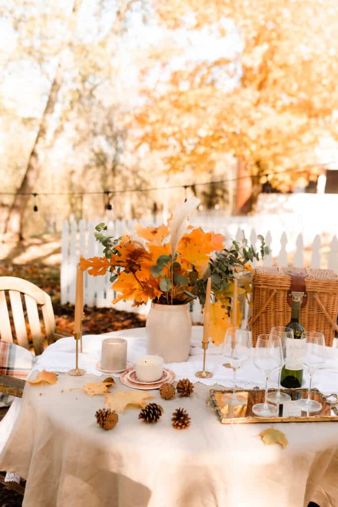 fall table setting outside on a farm. Table is covered with a tan linen table cloth, wine glasses on an antique mirrored tray, crock of dried leaves and eucalyptus and beeswax pillar candles in gold candlesticks