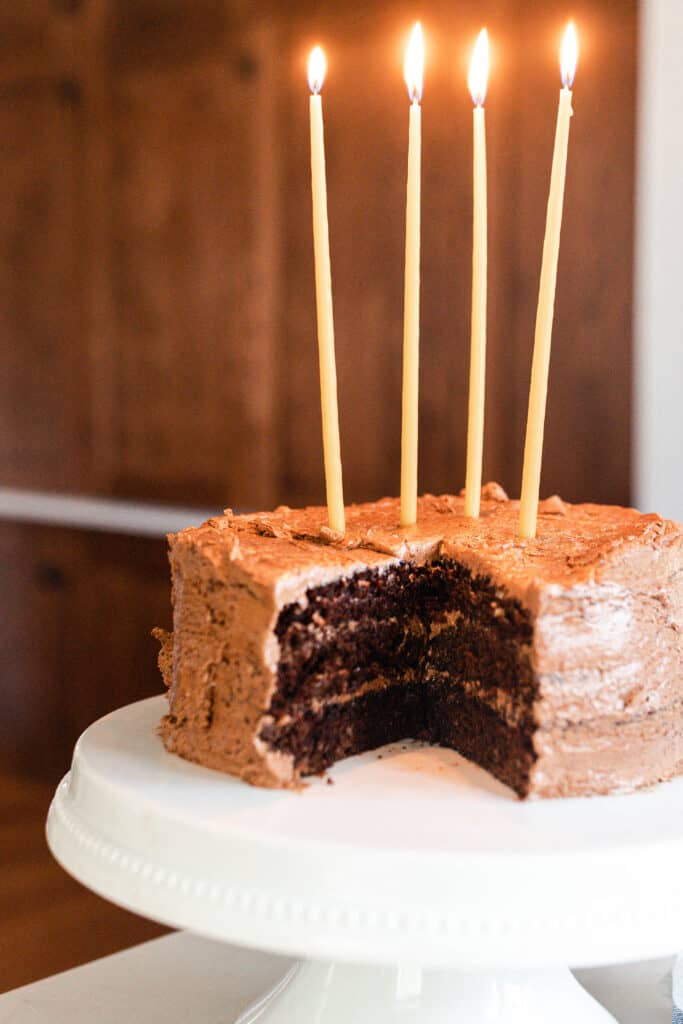 chocolate cake with chocolate frosting with 4 long candles on a white cake stand