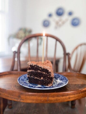 slice of three layer sourdough chocolate cake with chocolate frosting on a blue and white antique plate with a candle on a antique wooden high chair