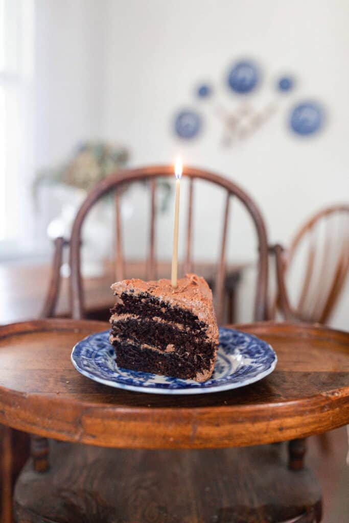 slice of three layer sourdough chocolate cake with chocolate frosting on a blue and white antique plate with a candle on a antique wooden high chair