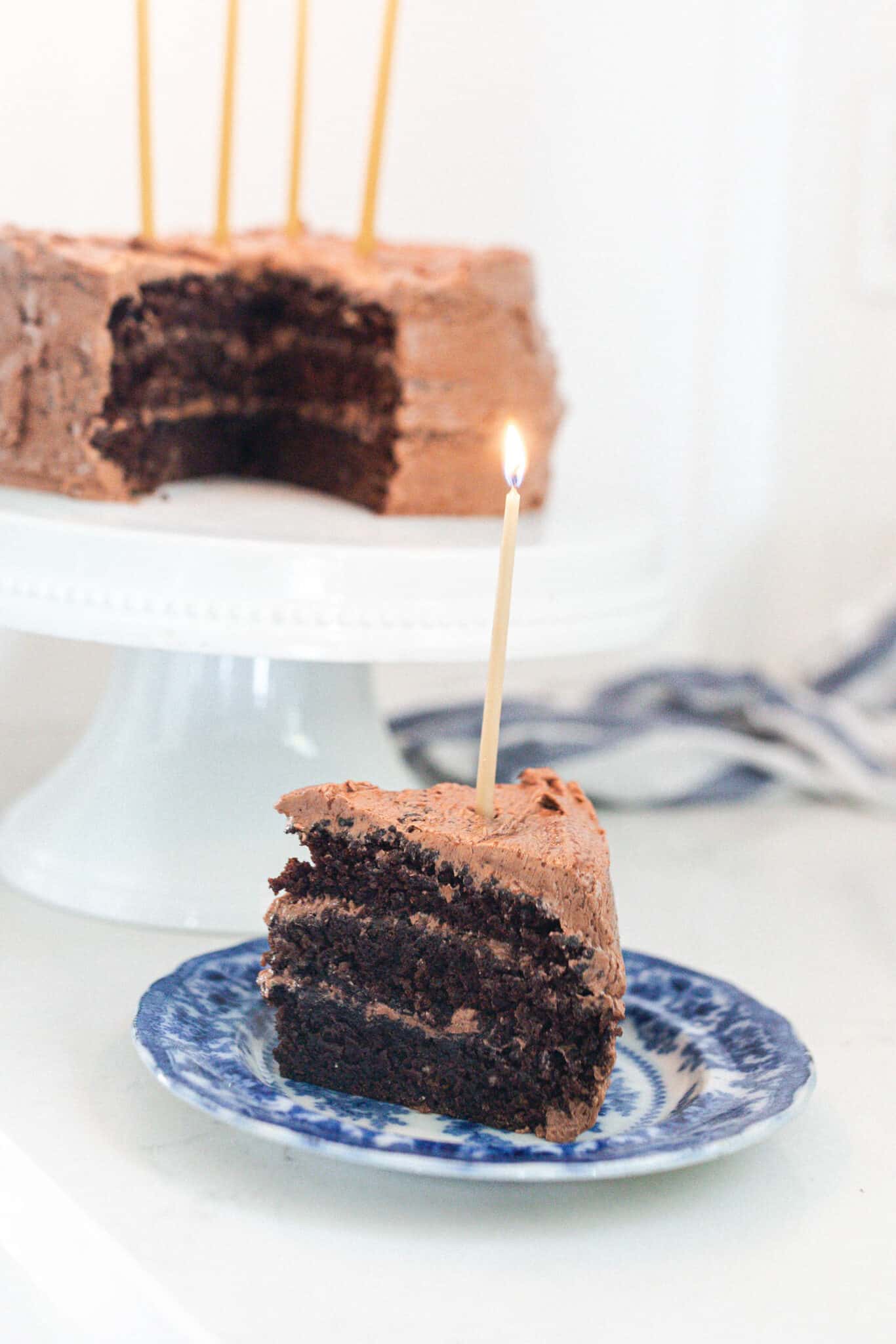 slice of three layer sourdough cake with chocolate frosting on a blue and white antique plate with the remaining cake on a white cake stand in the background