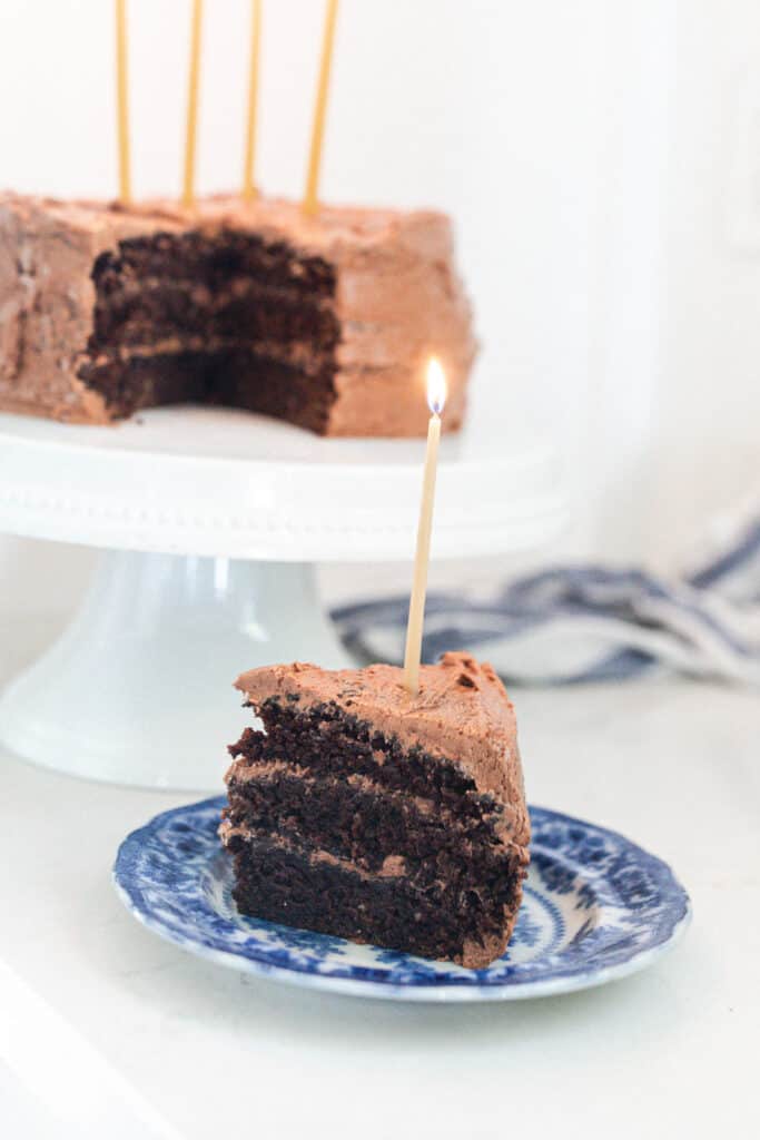 slice of three layer sourdough cake with chocolate frosting on a blue and white antique plate with the remaining cake on a white cake stand in the background