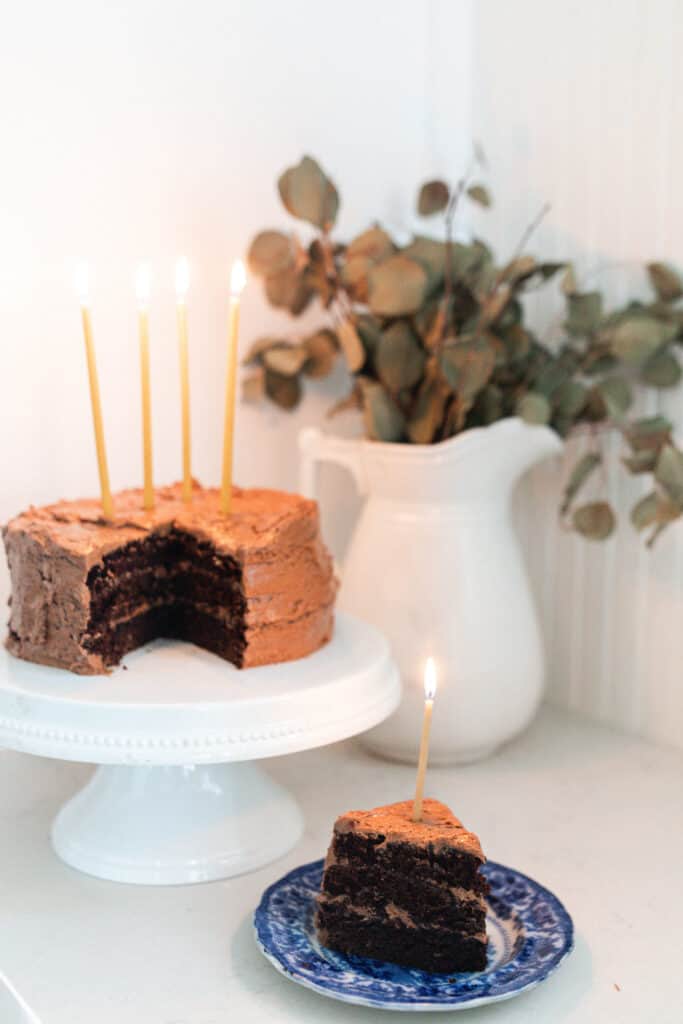 slice of sourdough discard chocolate cake on a blue and white antique plate with the rest of the cake on a white cake stand in the background with a case of eucalyptus