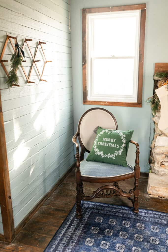 antique chair with a green Christmas pillow with a accordion coat hanger to the left with some greenery and bells hanging front it