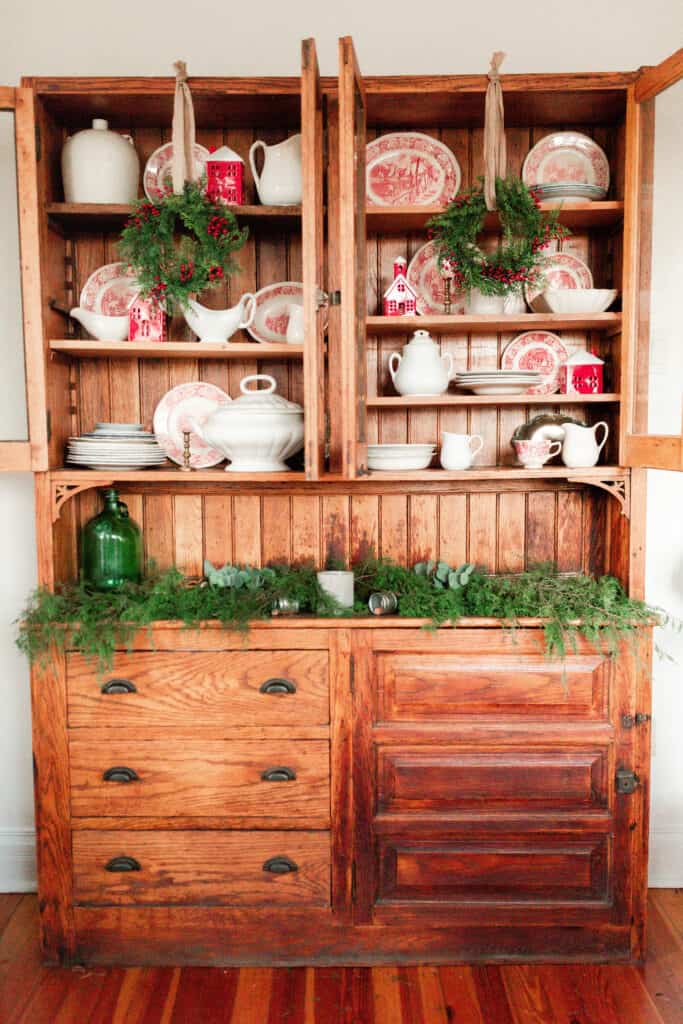 red and white plates, Christmas village houses and white vases and serving platter fill a wooden antique China cabinet. Green garland fill the cabinet countertop and wreaths hang with ribbon