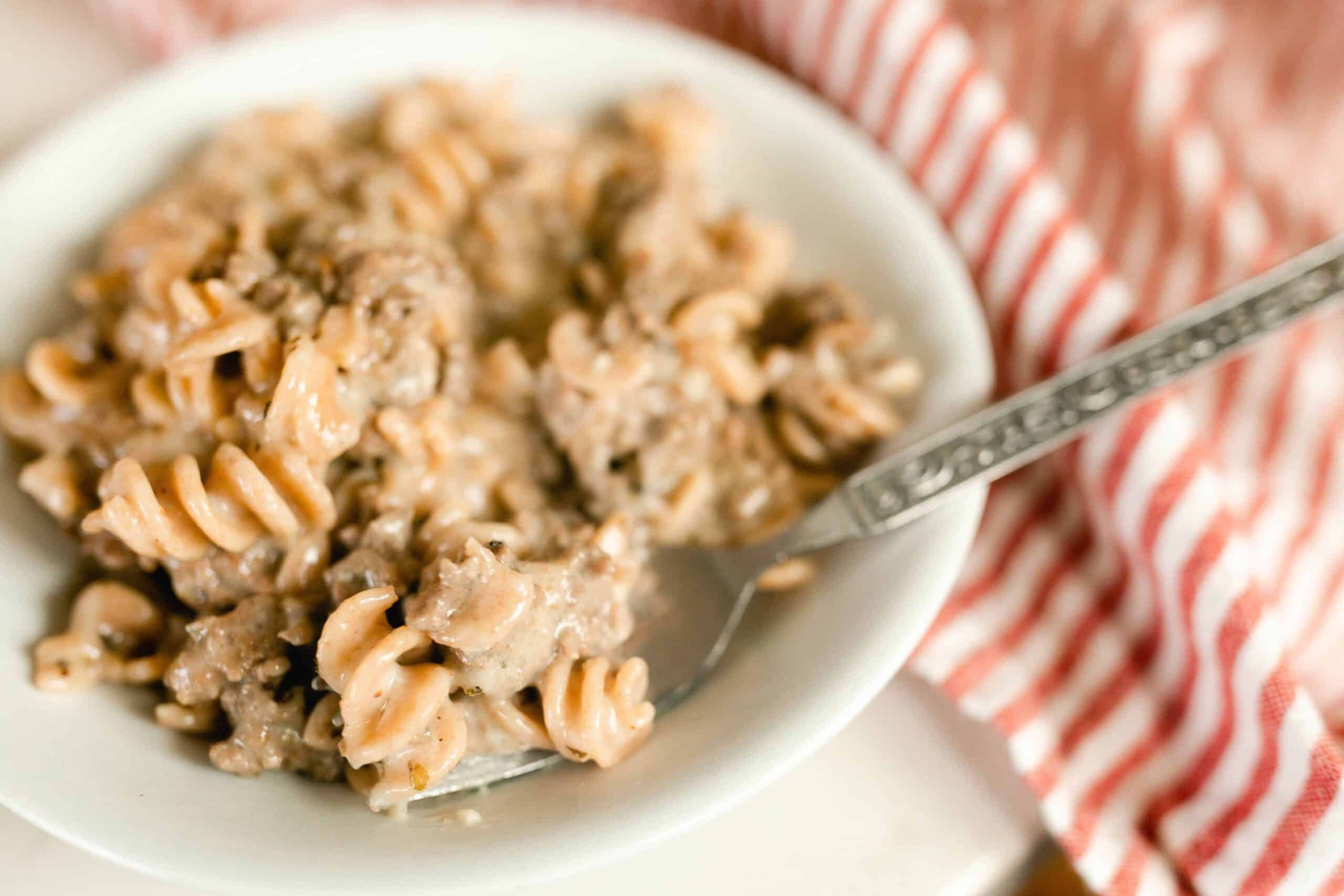 homemade hamburger helper in a white bowl with a fork in the pasta. a red and white stripped napkin to the right