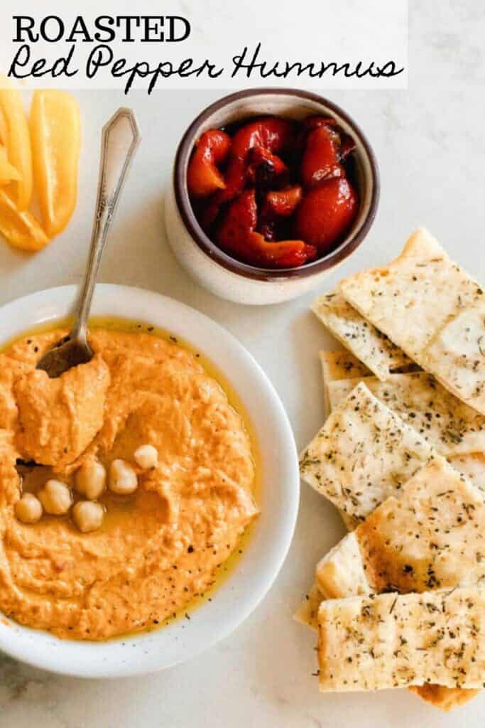 bowl of roasted red pepper hummus topped with chickpeas with a jar of roasted red peppers back in oil and sourdough flat bread to the right