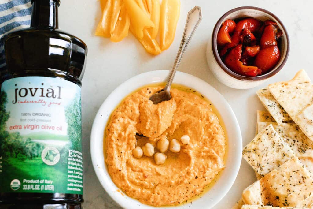 red pepper hummus in a bowl with roasted red peppers, olive oil, and sourdough flat bread surrounding the bowl