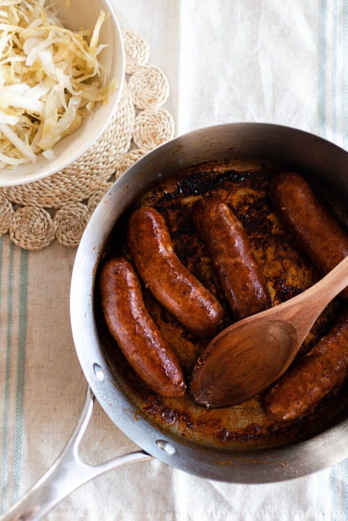 pan fried sausage in a skillet with a bowl of sauerkraut in the background