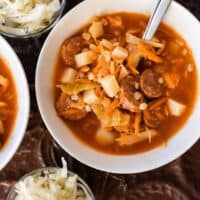 bowl of sauerkraut soup with sausages with a spoon in the bowl. Two glass bowls of sauerkraut sit next to the bowl of soup
