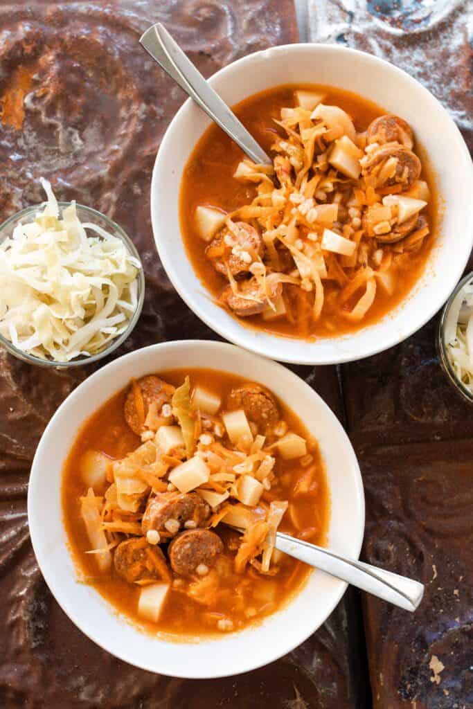 two bowls of sauerkraut soup with sausage on a brown tin table.