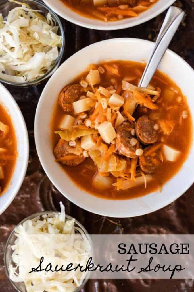 bowl of sauerkraut soup with sausages with a spoon in the bowl. Two glass bowls of sauerkraut sit next to the bowl of soup