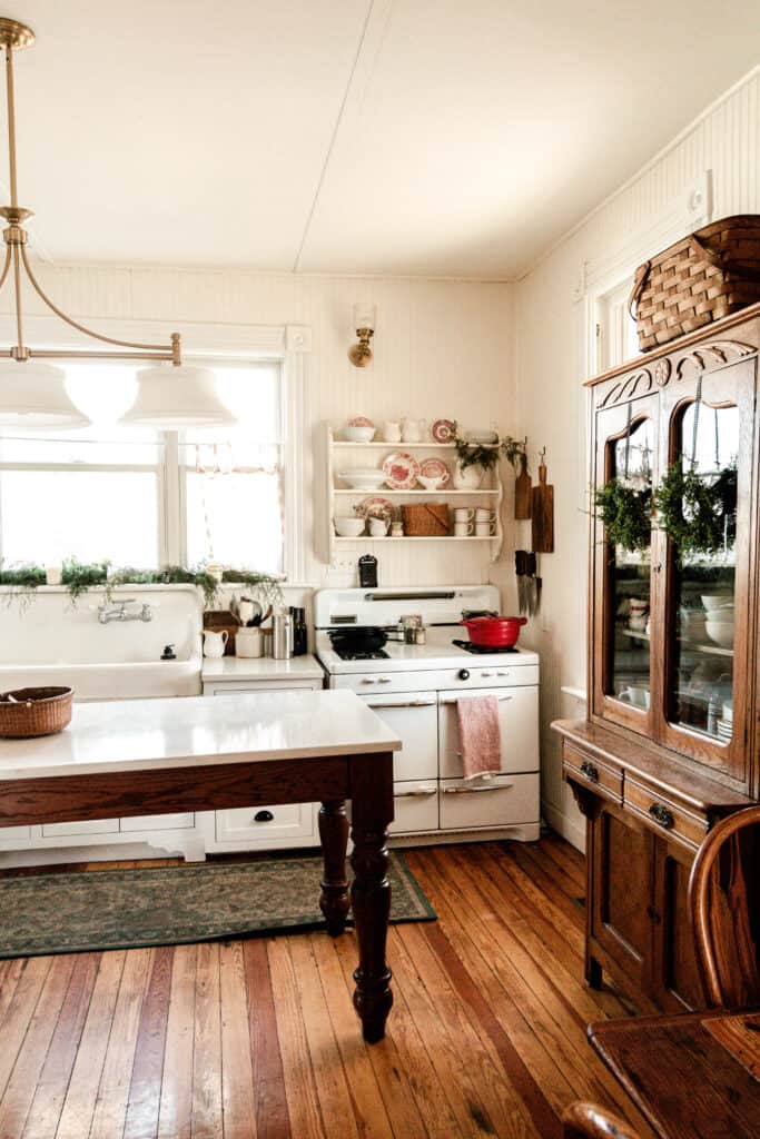 farmhouse kitchen decorated for Christmas with white and red plates sitting on the shelves over the stove. Greenery is layed on the window sill and wreaths hanging from china cabinet.