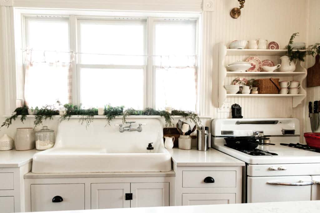 white farmhouse kitchen decorated for Christmas. Garland sits on the window sill, red and white plates and mugs are nestled on a an antique shelf