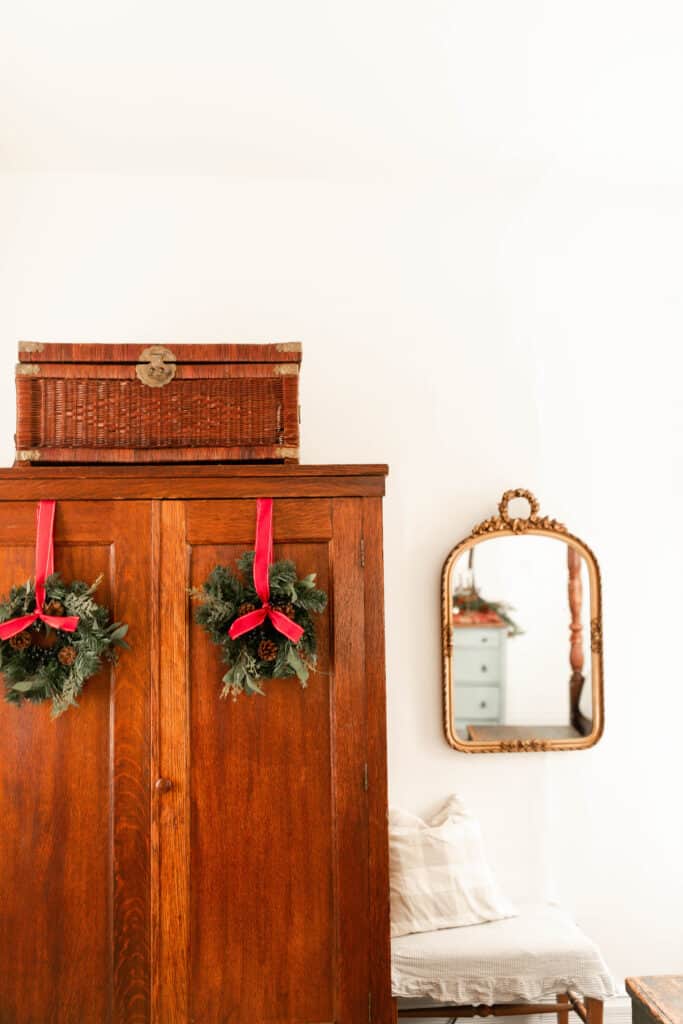 wood antique armoire with two greenery wreaths hanging with red ribbon. A gold mirror hangs to the right above a vintage bench