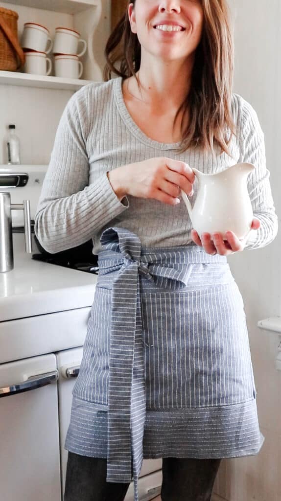 women wearing a gray sweater and a blue and white half apron holding a white vase in front of a vintage stove.