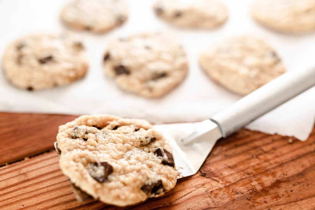 spatula with a sourdough chocolate chip cookie on it. More cookies in the background on parchment paper.