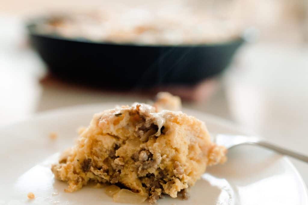 slice of breakfast strata on a white plate with a fork. A cast iron skillet is in the background