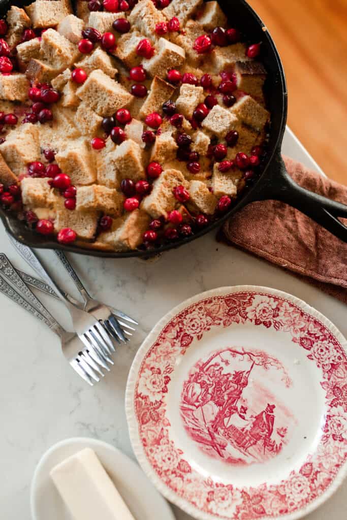 a antique red and white plate with forks and a cast iron skillet in the background with French toast casserole and cranberries.