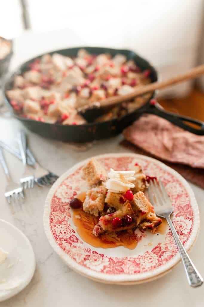 cranberry French toast casserole on a red and white antique plate with a fork. A cast iron skillet with more casserole in the background.
