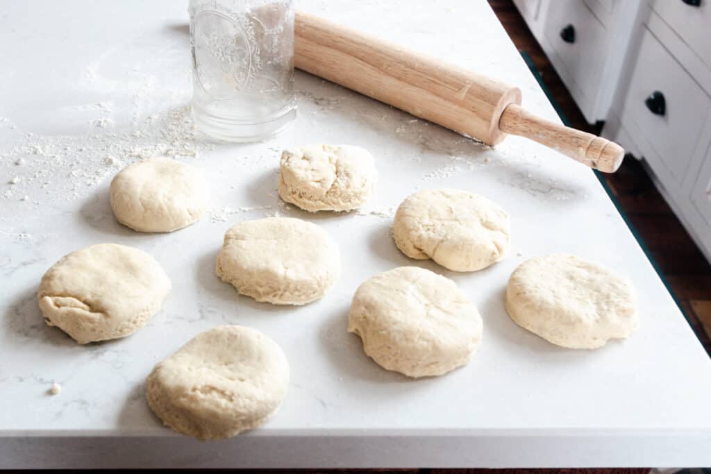 einkorn biscuits cut out and laying on a white quartz countertop with a rolling pin and a mason jar in the background