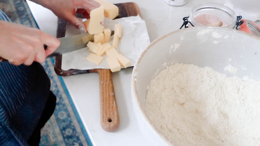 women chopping butter on a small wooden cutting board and adding it to a bowl of dry ingredients