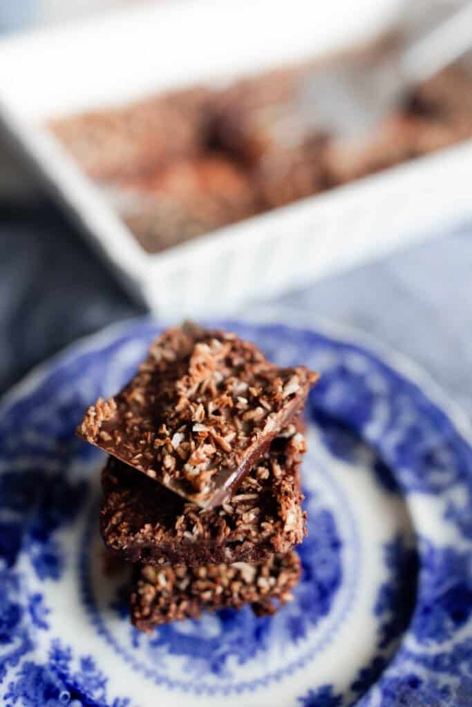 chocolate superfood bars squares topped with toasted coconut stacked on a antique white and blue plate with a baking dish of more bars