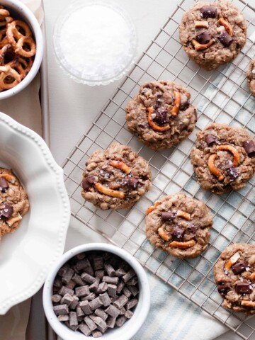 overhead photo of a cooling rack of chocolate oatmeal lactation cookies with a plate of cookies to the left and two white dishes of pretzels and chunks