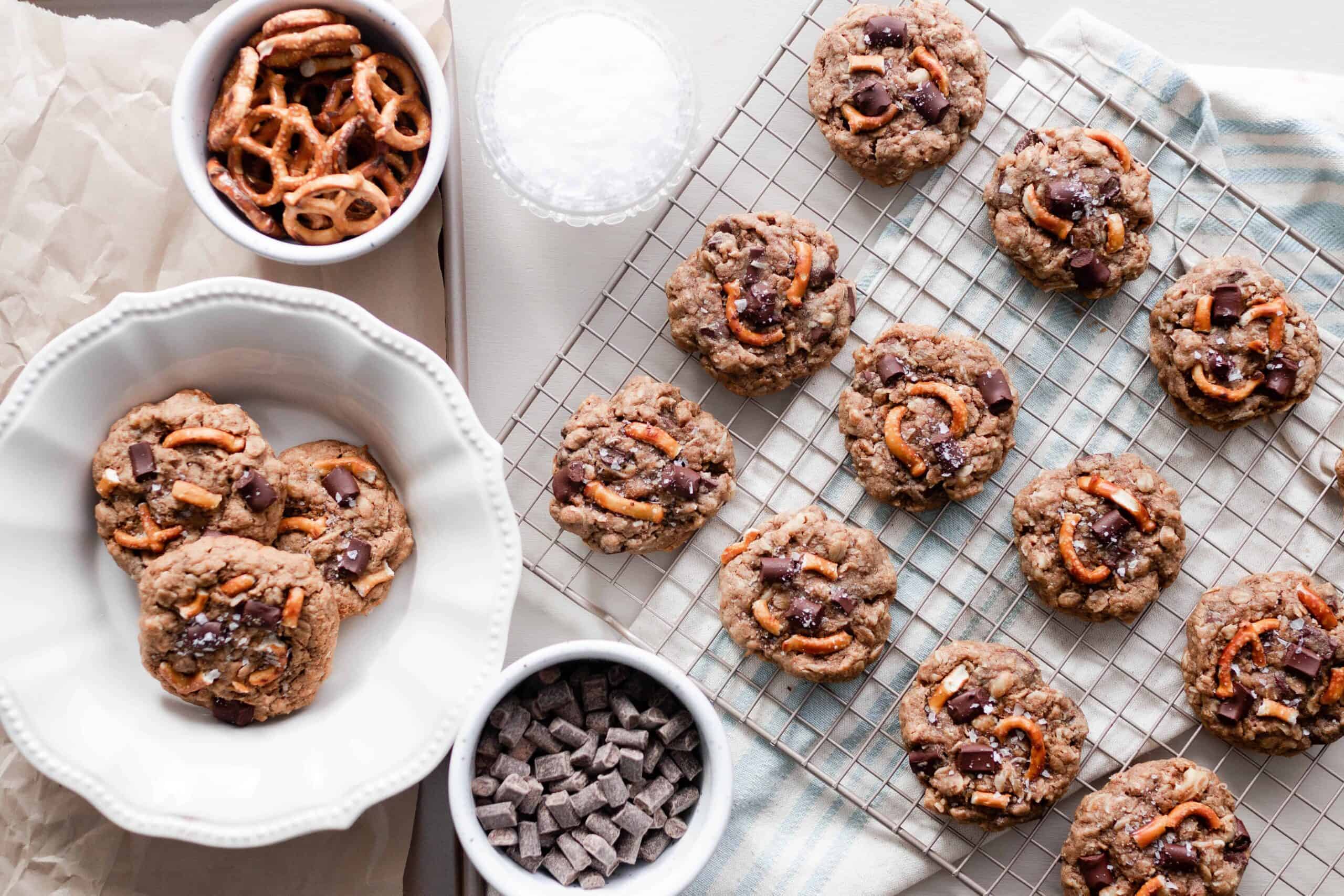 overhead photo of a cooling rack of chocolate oatmeal lactation cookies with a plate of cookies to the left and two white dishes of pretzels and chunks