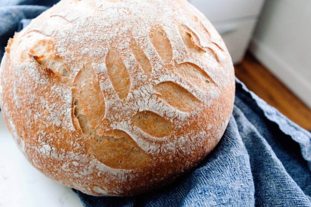 no knead sourdough bread loaf with leaf design on a white countertop with a dark blue towel